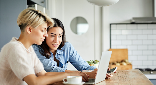 Deux femmes utilisent un ordinateur portable. L’une d’elles, portant un haut blanc, tape sur les touches du clavier, tandis que l’autre, portant un haut bleu, regarde l’écran de l’ordinateur un smartphone à la main.