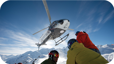 Imagen de un helicóptero de heliesquí sobrevolando un grupo de esquiadores en una cumbre de montaña.